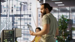Cheerful admin holding laptop chatting with colleagues while working with machine learning tech. Man transforming data into predictive models using notebook and talking with coworkers, camera A